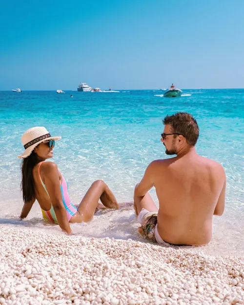 Paar entspannt am türkisfarbenen Strand von Sardinien mit kristallklarem Wasser und Segelbooten im Hintergrund